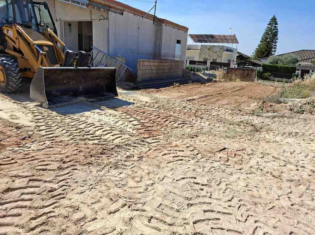 Obra en construcción con excavadora amarilla, terreno de tierra y un almacén bajo un cielo azul despejado.