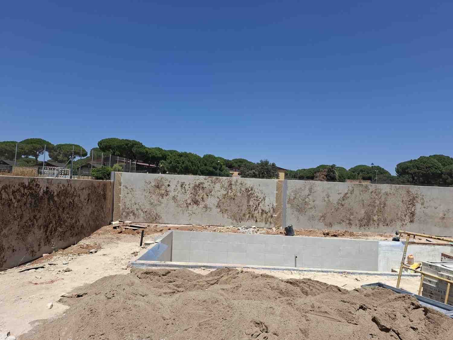 Obra en construcción con foso de cimentación de hormigón, terreno de tierra y muros de piedra bajo un cielo azul despejado.