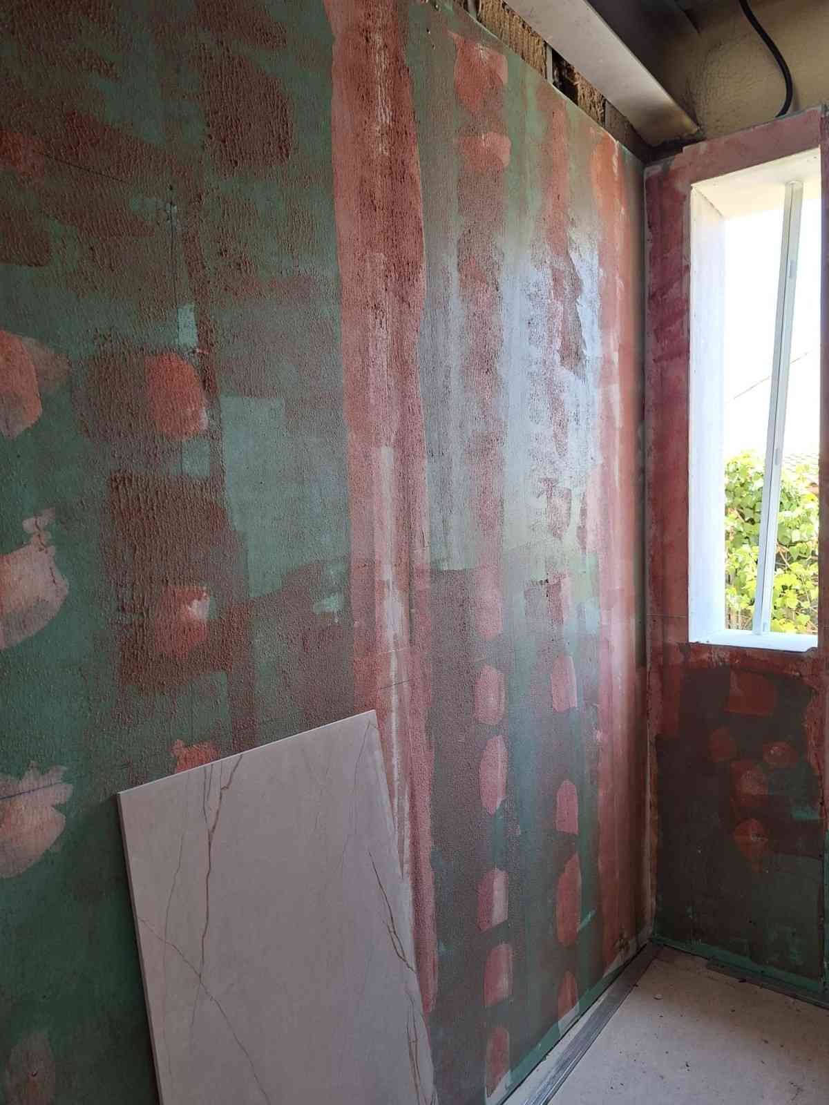 Habitación sin terminar con paredes de yeso verde y rojo, una ventana y una pizarra blanca apoyada contra la pared.