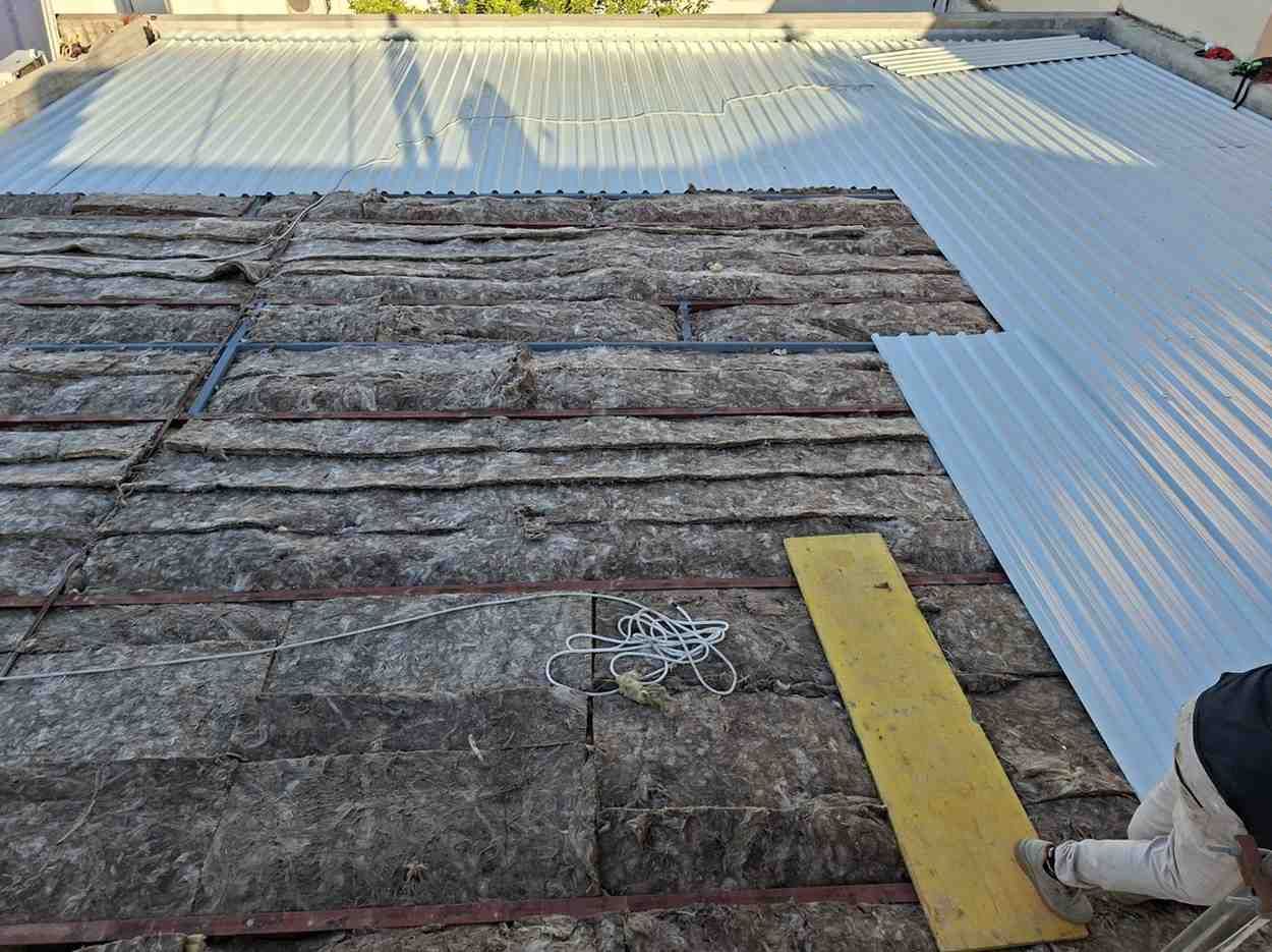 Techo en reparación con láminas de asfalto enrolladas y paneles de metal blanco; se ve la bota de un trabajador en el borde.