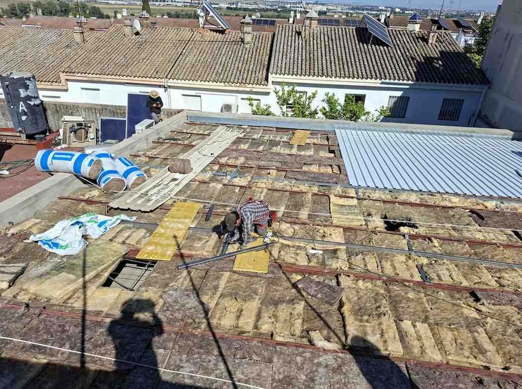 Azotea en reparación con techo expuesto, trabajadores, lonas azules y casas vecinas al fondo.