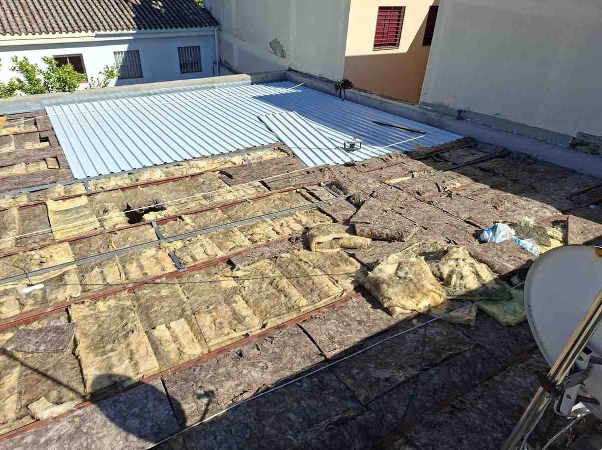 Azotea con aislamiento parcialmente retirado y tablas expuestas junto a un panel de techo de metal azul.