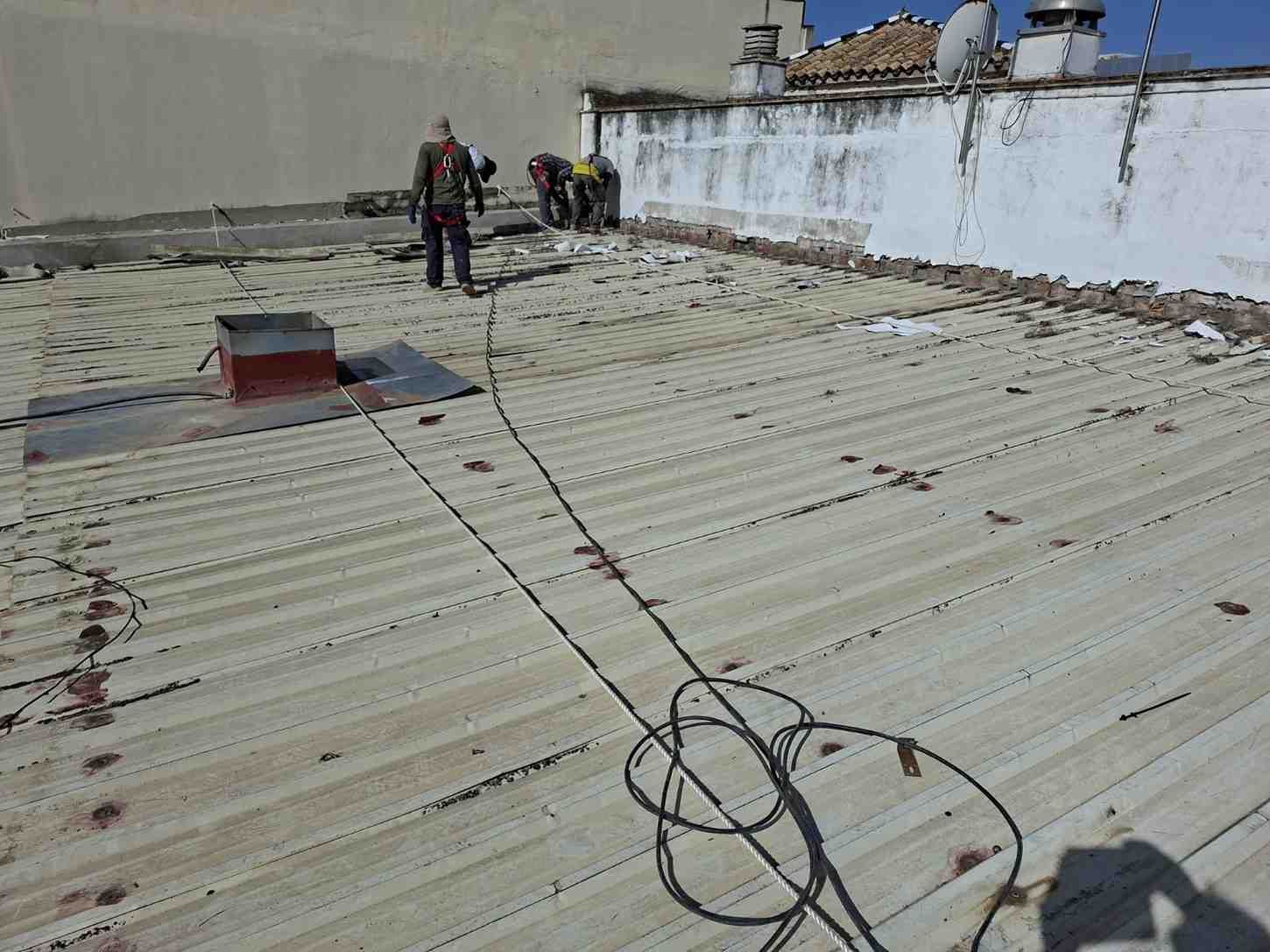 Trabajadores en un tejado de chapa ondulada con cables, cerca de una pared blanca bajo un cielo despejado.