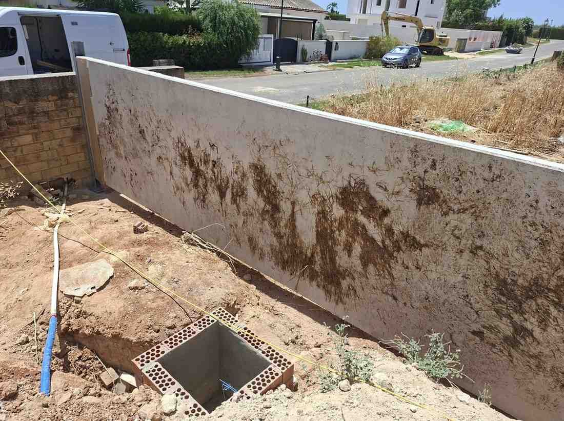 Muro de hormigón en un patio polvoriento con una pequeña abertura cuadrada para servicios públicos en el suelo.