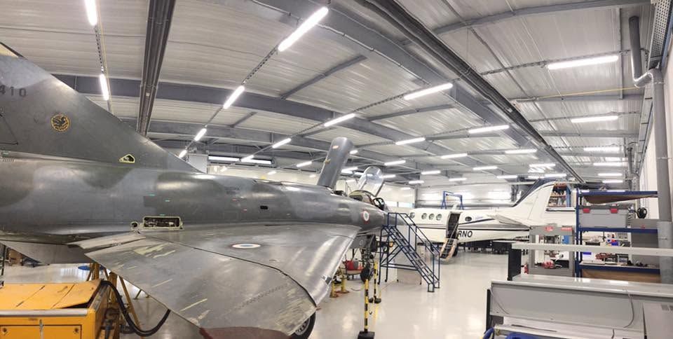 Un avion de chasse à l'intérieur d'un hangar, éclairé par des projecteurs puissants, avec des établis à proximité.