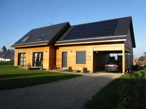 Maison moderne en bois avec panneaux solaires, allée privée et voiture garée dans le garage.