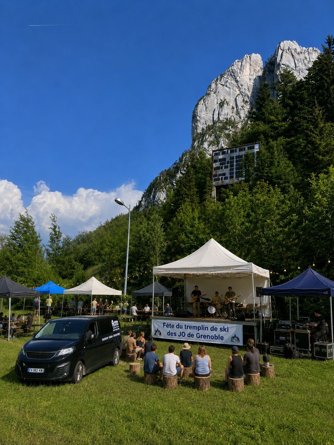 Scène extérieure avec des orateurs et un public sous des tentes à côté d'une voiture, au pied d'une falaise rocheuse.
