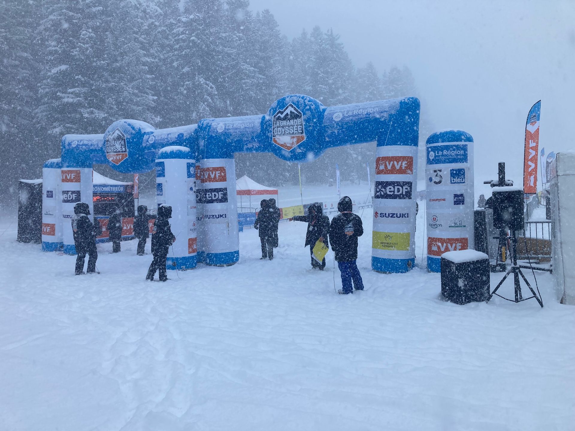 Entrée enneigée d'une piste de ski en plein air avec une arche, des bannières de sponsors et des personnes debout dans une épaisse couche de neige.
