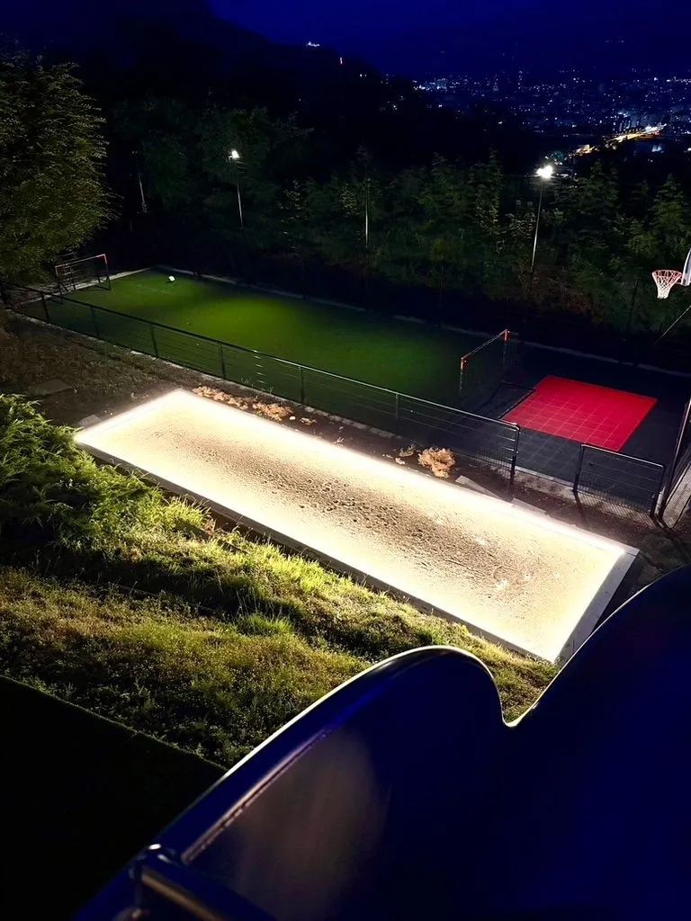 Vue nocturne d'un court de tennis éclairé avec un carré de service, entouré d'arbres et de tribunes.