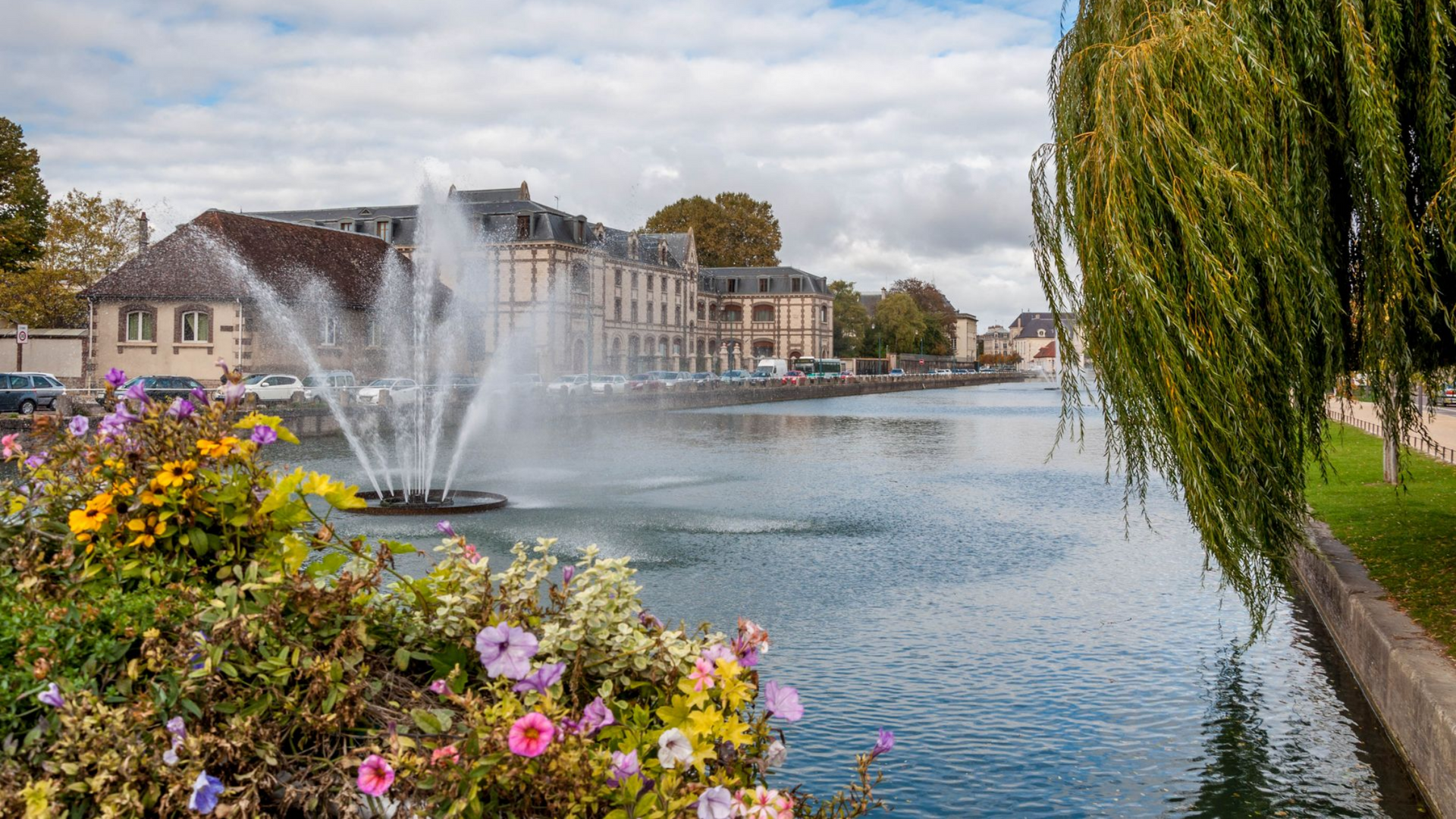 Fontaine dans un canal avec des fleurs et des bâtiments, sous un ciel nuageux.