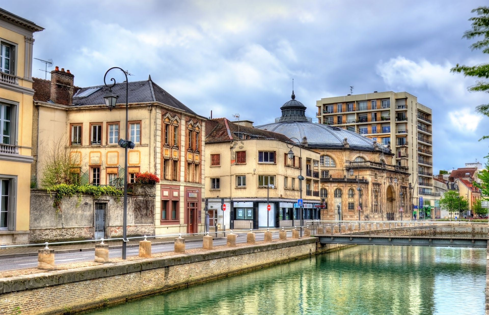 Bâtiments bordant un canal dans une ville européenne. Architecture en pierre beige, ciel nuageux, reflets dans l'eau.