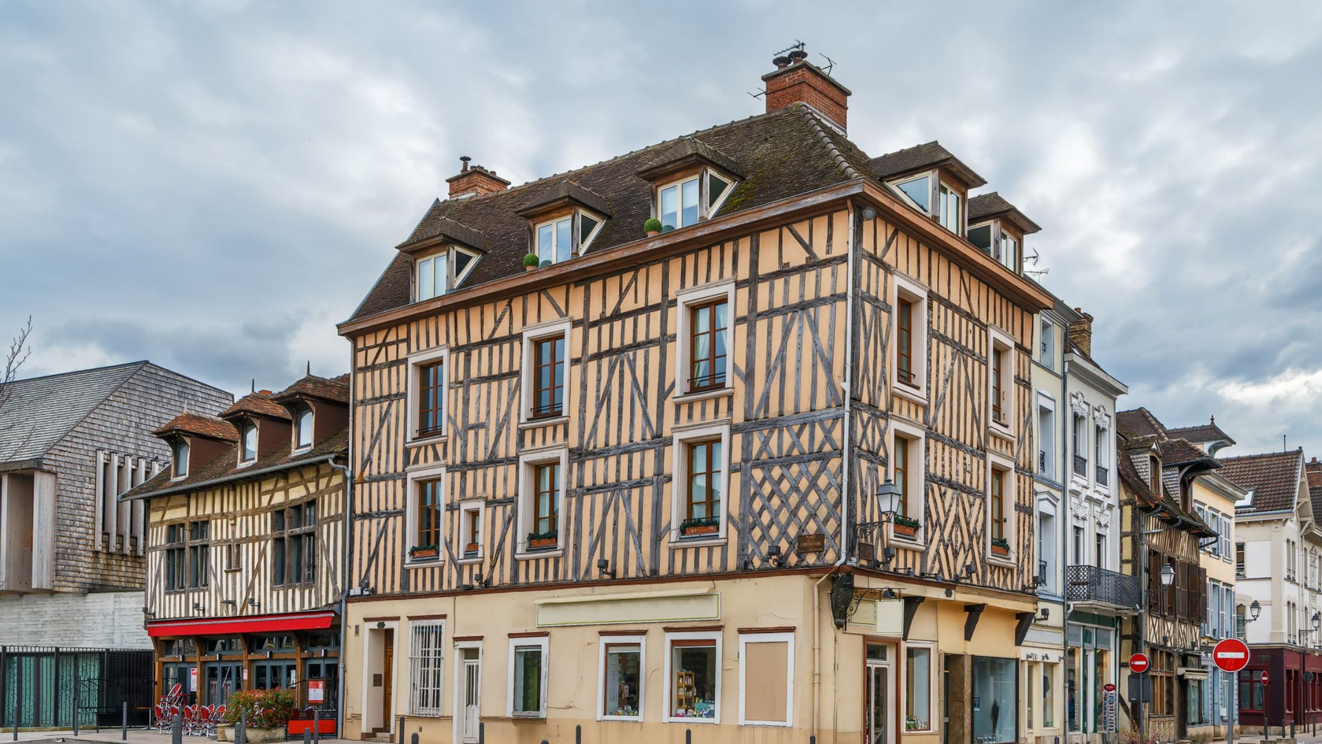 Bâtiment à colombages au coin d'une rue, murs beiges, poutres en bois foncé, toit marron, ciel nuageux.