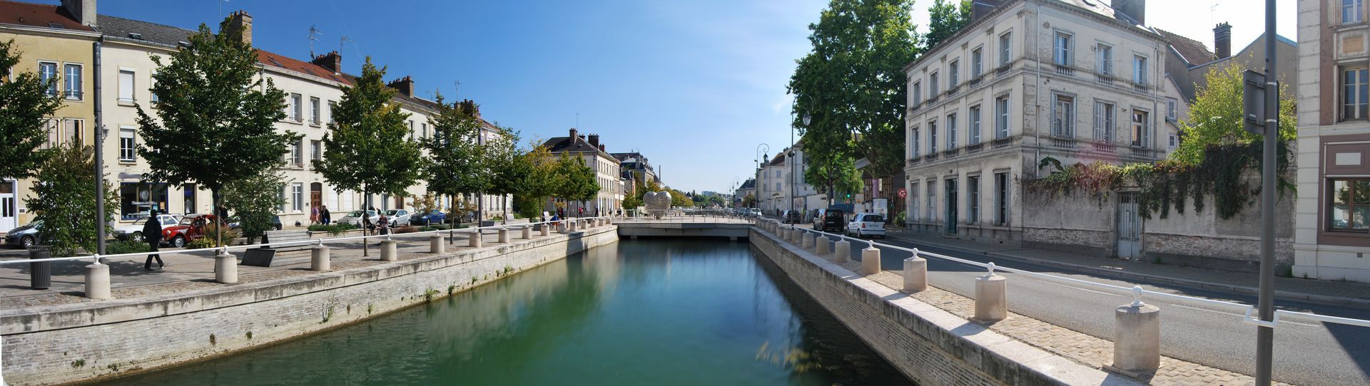 Un canal traverse une ville européenne bordée de bâtiments et d'arbres. Ciel bleu.