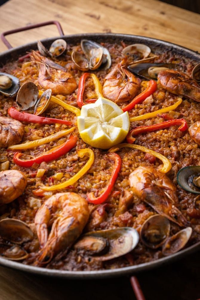 Una paella con camarones, mejillones, pimientos y un limón sobre una mesa de madera.