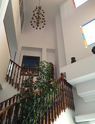 Escalera de madera con barandilla, planta, obra de arte y lámpara de araña en un interior luminoso.