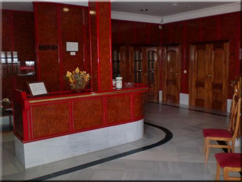 Mostrador de recepción de hotel con detalles en rojo y marrón, arreglo floral y puertas de madera cerradas.