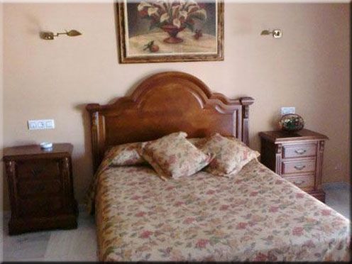 Dormitorio con cama de madera, ropa de cama floral, dos mesitas de noche, cuadro y apliques de pared.