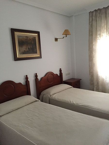 Dos camas individuales en una habitación con un cuadro, una lámpara de pared y una ventana con cortinas transparentes.