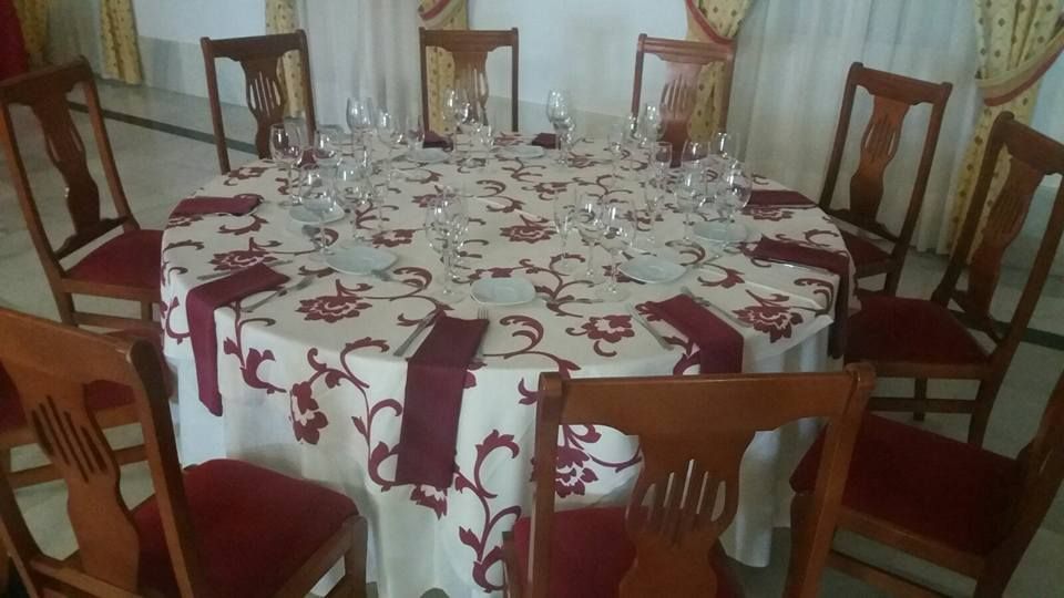 Mesa redonda para comer con ocho sillas de madera. Mantel blanco, diseño floral rojo y cristalería.