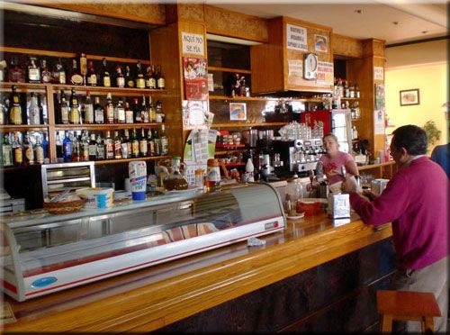 Interior del bar: Una persona pide en la barra. Estantes llenos de botellas detrás; una mujer prepara una bebida.