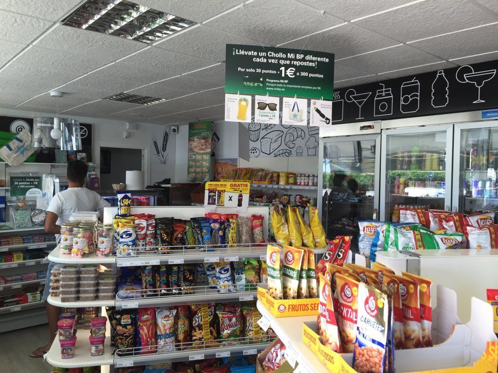 Interior de una tienda de conveniencia con estantes de bocadillos, persona en el mostrador y refrigerador.