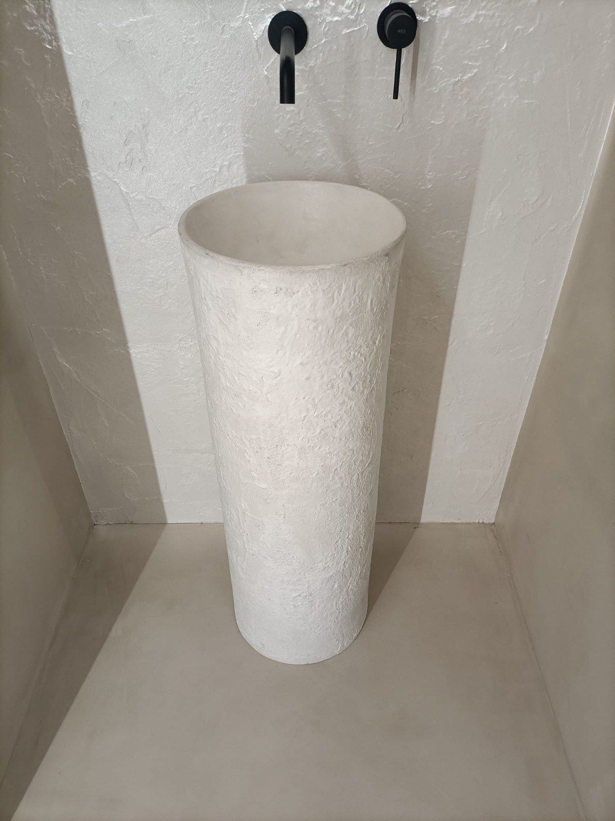 Lavabo de pedestal blanco con grifos negros en un pequeño nicho texturizado.