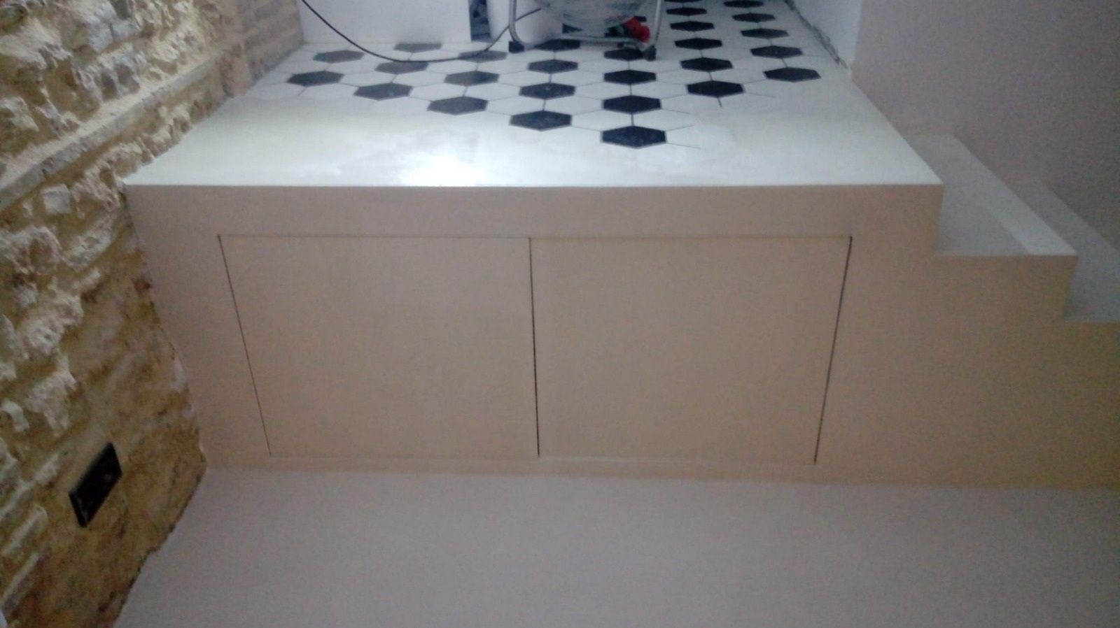 Plataforma de almacenamiento blanca con dos puertas y suelo de baldosas estampadas. Pared de piedra visible.