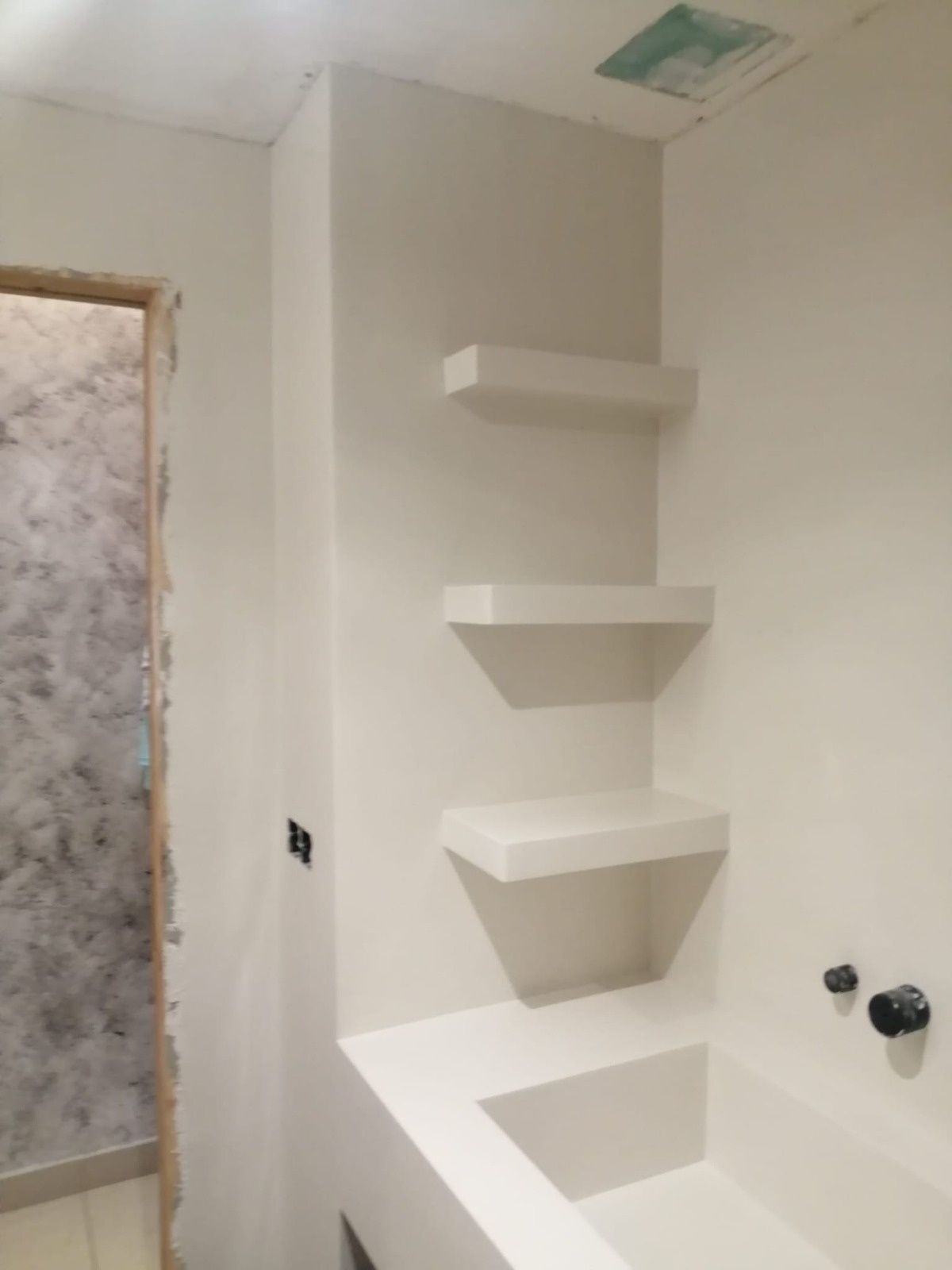 Baño esquinero blanco con tres estantes empotrados sobre una bañera empotrada. Se ven la puerta y los sanitarios.