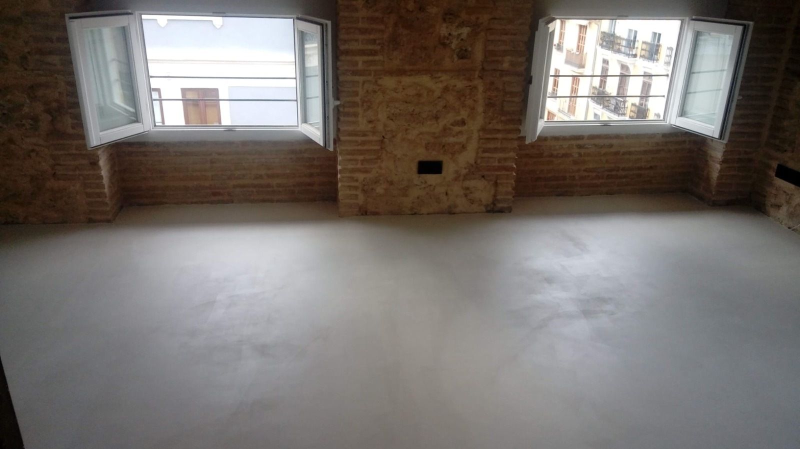 Habitación vacía con suelo de color claro, paredes de ladrillo y dos ventanas abiertas.