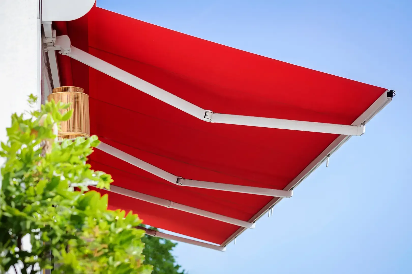 Eine leuchtend rote, ausfahrbare Markise erstreckt sich von einer weißen Wand neben einer hellen Laterne vor einem klaren blauen Himmel.