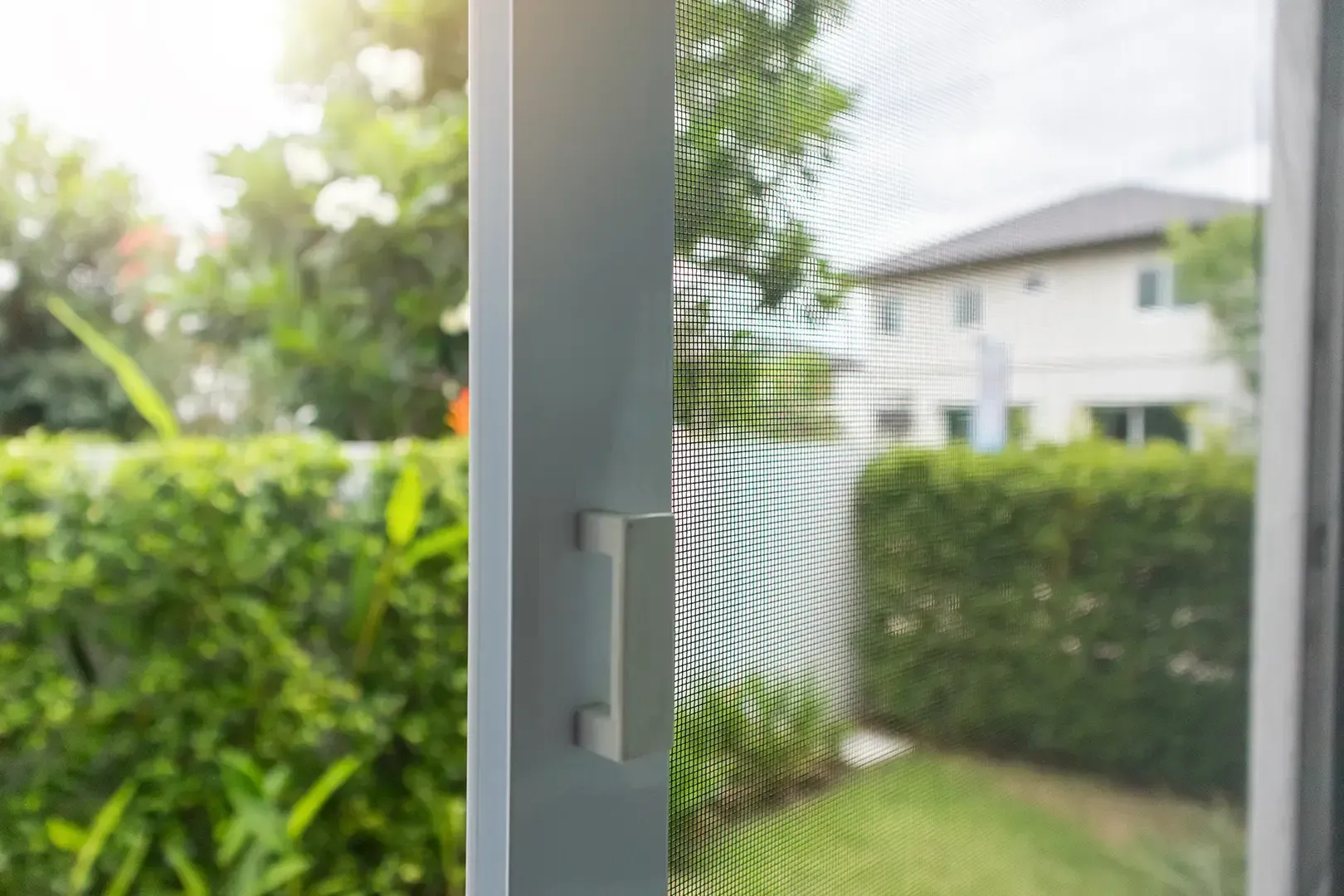 Nahaufnahme eines weißen Fliegengittertürgriffs mit verschwommener Ansicht eines sonnigen grünen Gartens und eines Hauses im Hintergrund.