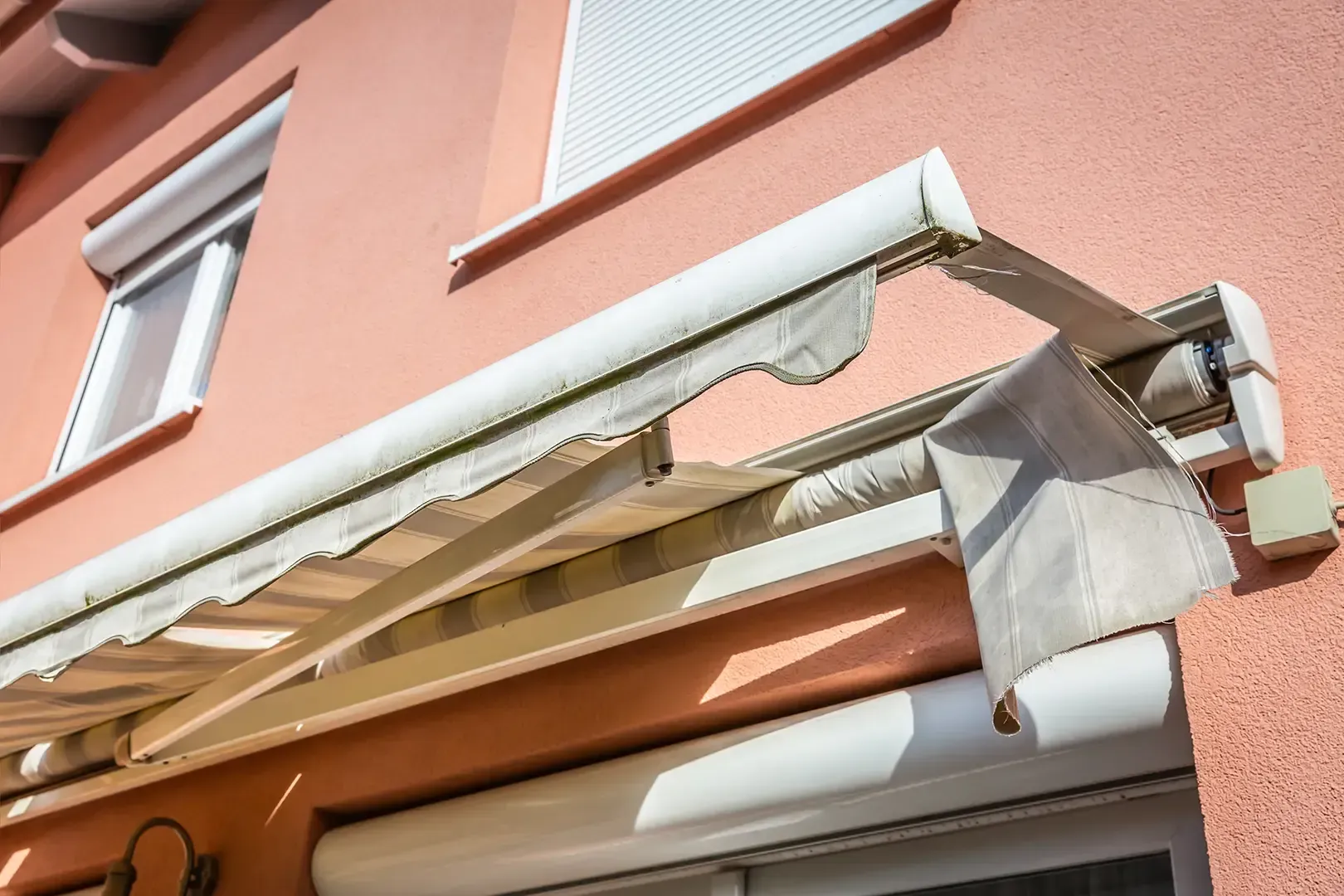 Eine ausladende, beige-weiß gestreifte und zerrissene Markise, die an einer lachsfarbenen Hauswand über einem Fenster befestigt ist.