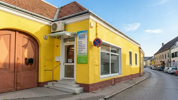 Ein gelbes, eingeschossiges Eckgebäude mit weißem Eingang und großen Fenstern, gelegen an einer Straße mit geparkten Autos.