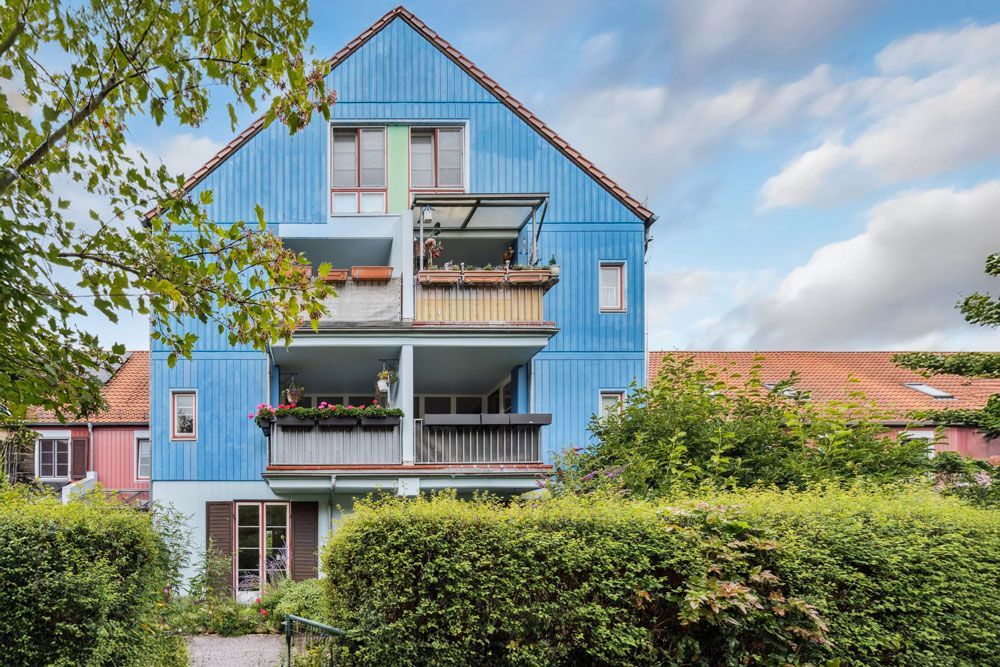 Blaues, mehrstöckiges Holzgebäude mit Balkonen, umgeben von grünen Büschen und Bäumen unter einem bewölkten Himmel.