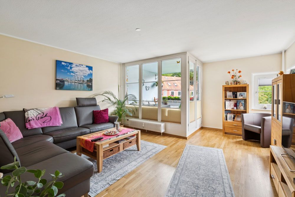 Wohnzimmer mit dunkelgrauer Eckcouch, Holzboden, großen Fenstern und einem Teppich.