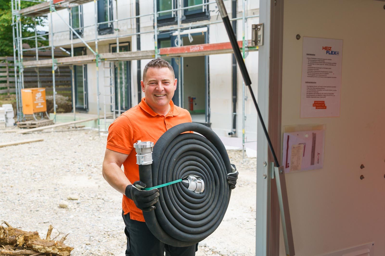 Ein lächelnder Arbeiter in orangefarbenem Shirt hält einen aufgerollten schwarzen Schlauch mit Metallkupplung.