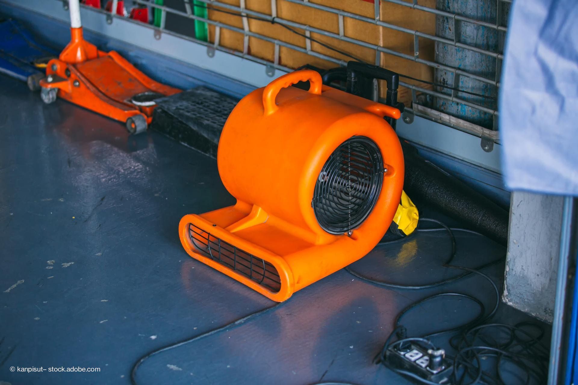Ein orangefarbener Bodenventilator steht in einer Werkstatt auf dem Boden, daneben liegen Kabel und Werkzeuge.
