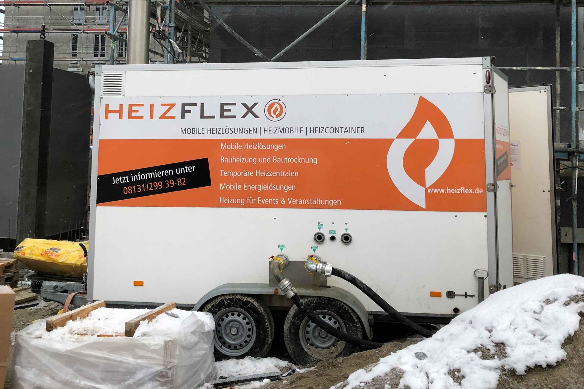 Ein weißer HeizFlex-Anhänger mit Anschlüssen und Schläuchen steht auf einer Baustelle, beworben als mobile Heizlösung.