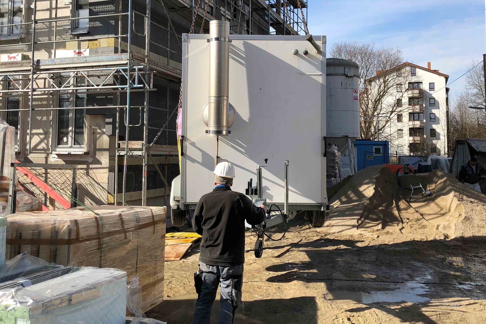 Ein Bauarbeiter mit Schutzhelm steht vor einem weißen Heizcontainer-Anhänger mit Abgasrohr auf einer Baustelle.