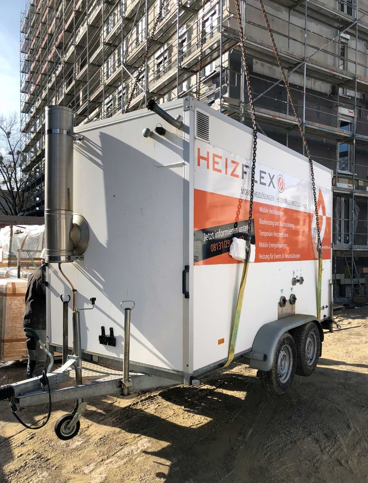 Ein HeizFlex-Anhänger mit Abgasrohr wird per Kettenzug auf einer Baustelle angehoben.