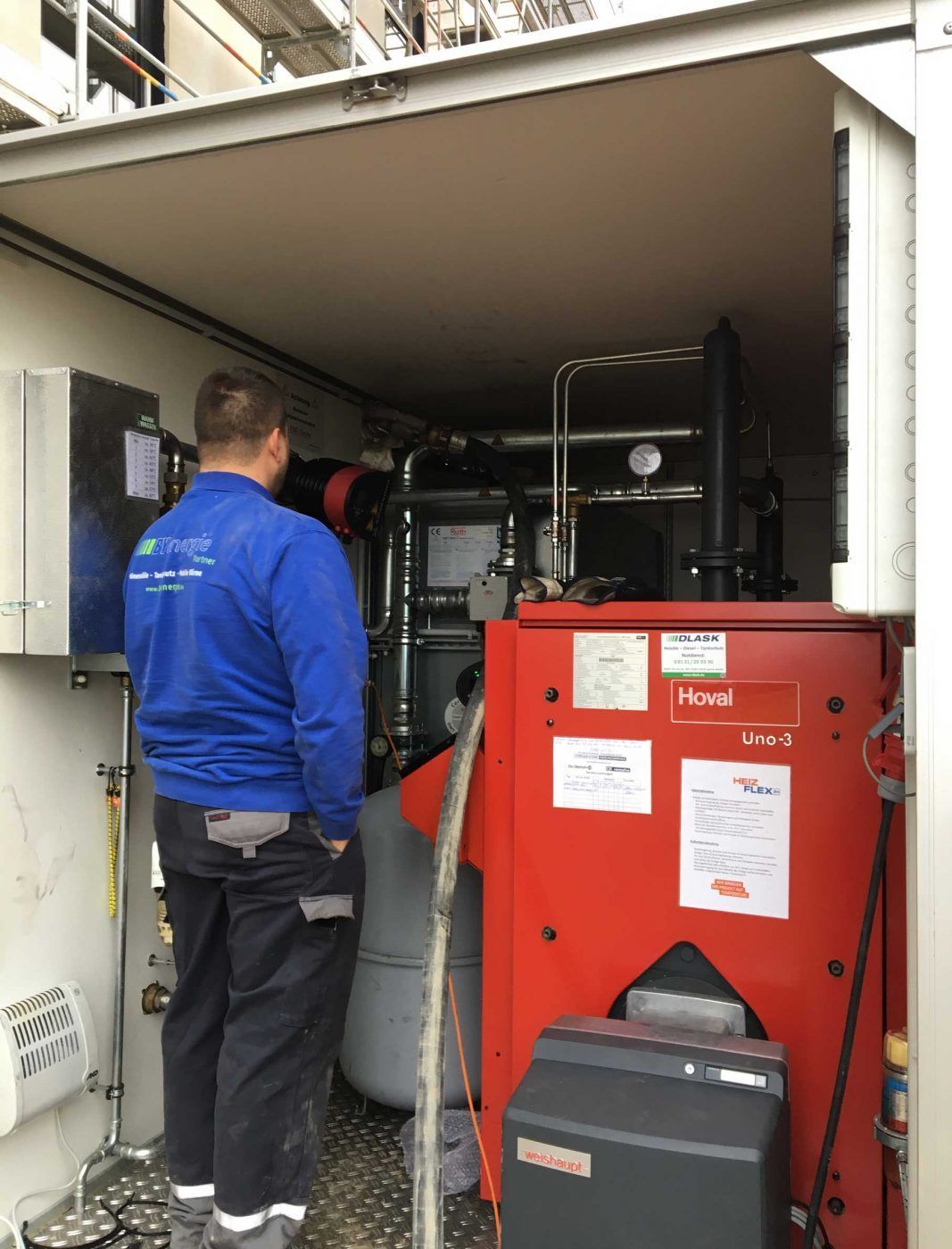Ein Arbeiter in blauer Jacke steht in einem Anhänger vor einer großen roten Heizungsanlage mit Rohren und Messgeräten.