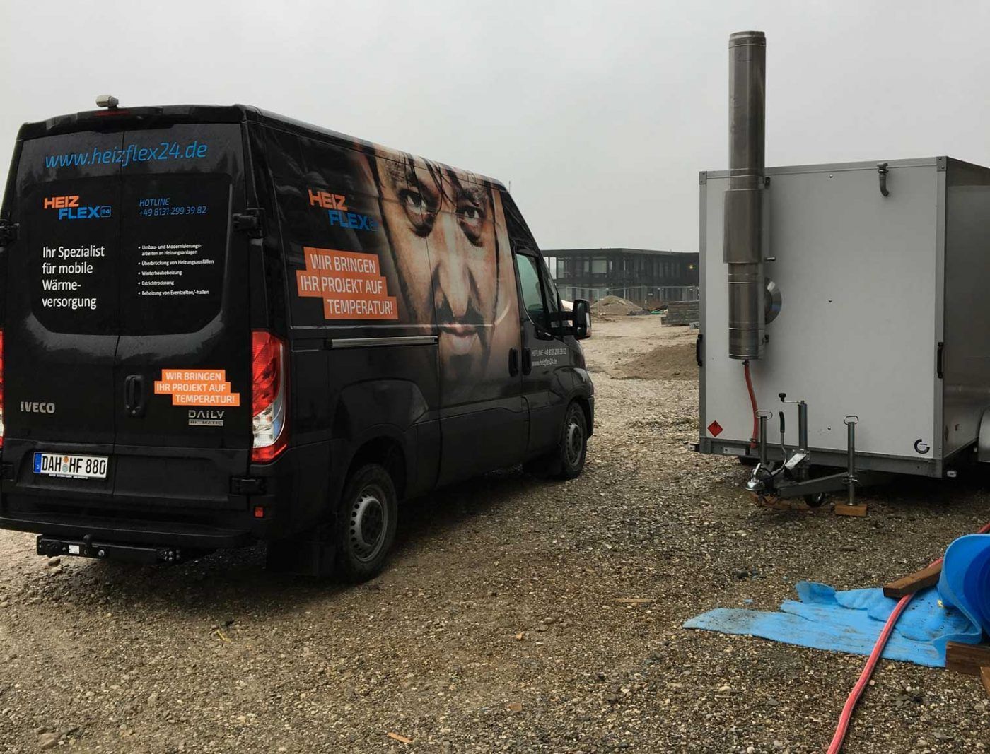 Ein schwarzer Transporter der Firma „HeizFlex24“ steht neben einem weißen Anhänger mit Abgasrohr auf einer Baustelle.