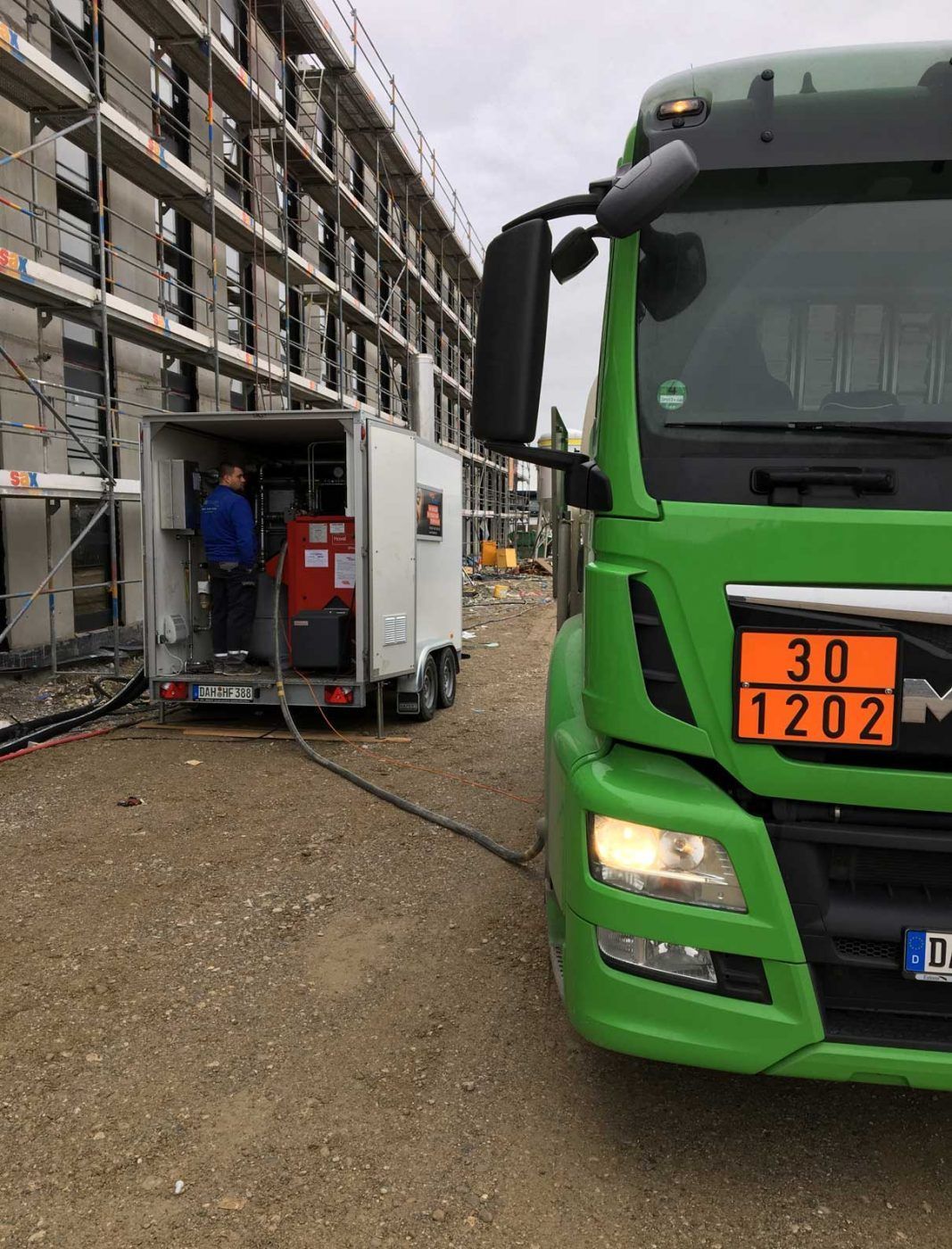 Ein grüner Lkw  steht auf einer Baustelle, verbunden über Schläuche mit einem weißen Anhänger, in dem ein Arbeiter arbeitet.