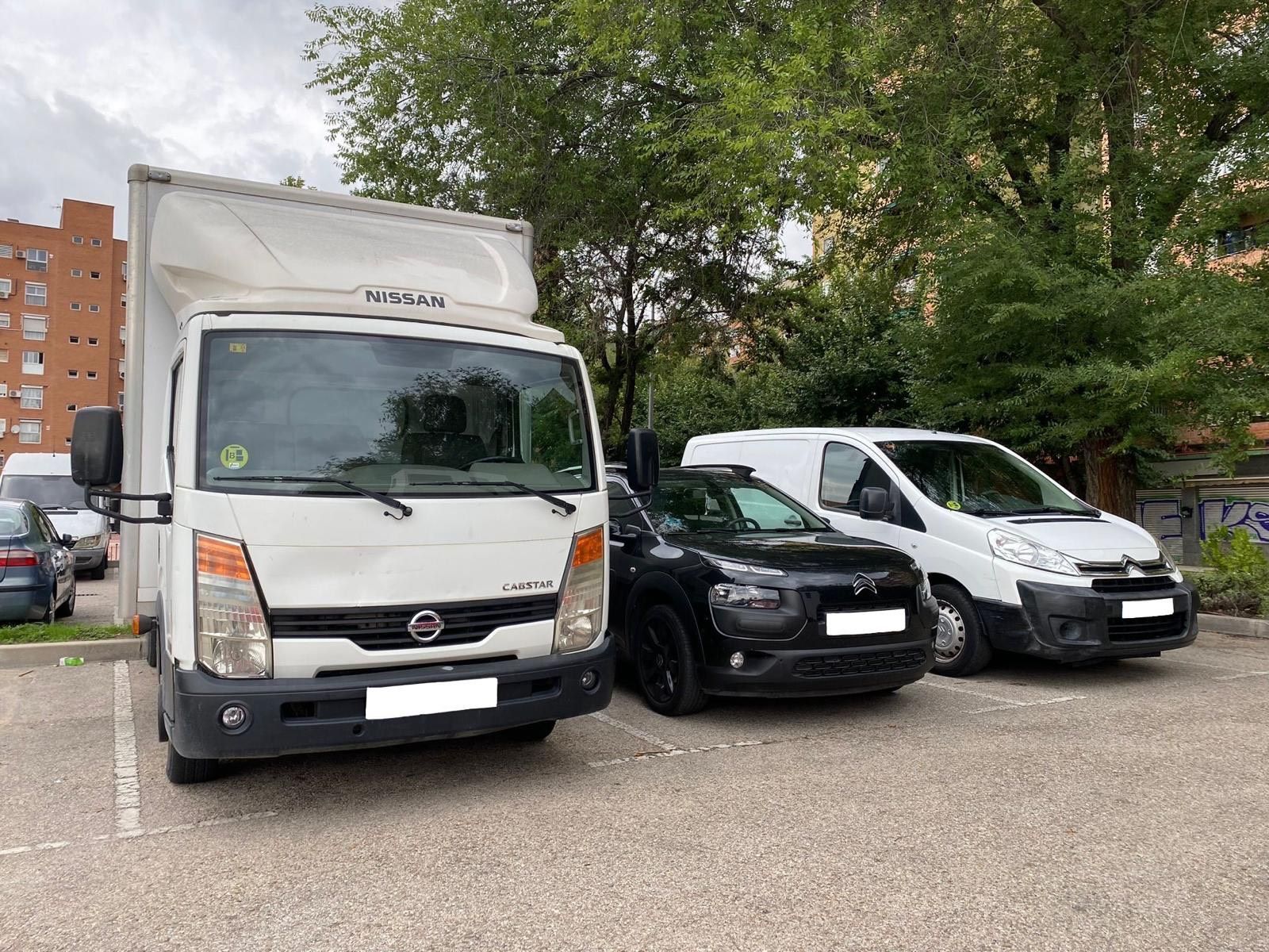 Un camión blanco, un automóvil negro y una camioneta blanca estacionados en un lote, con árboles y un edificio al fondo.