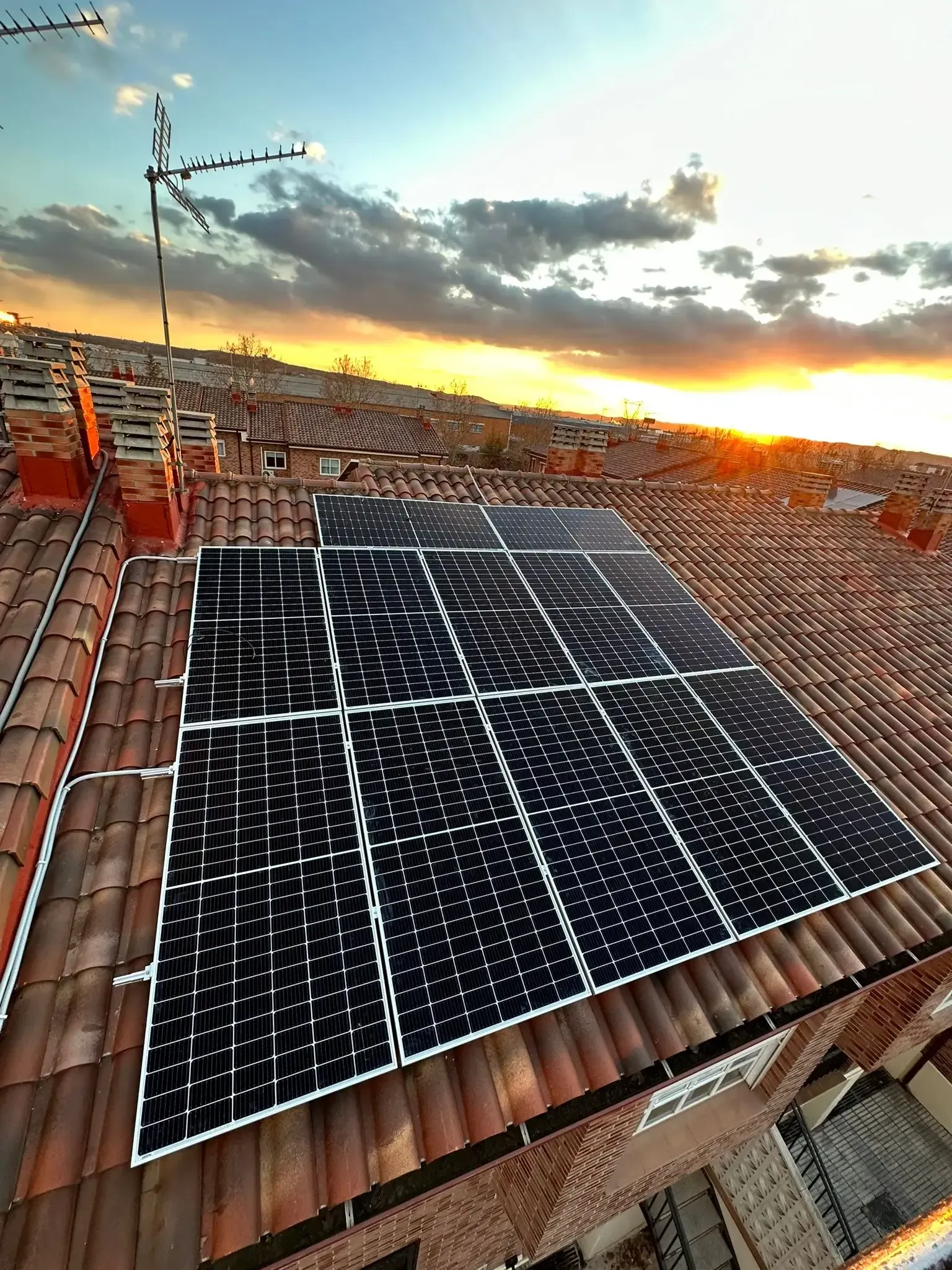 Paneles solares en un tejado de tejas rojas al atardecer, con nubes y un cielo anaranjado cálido.