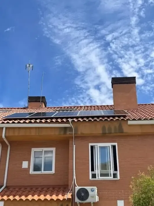 Fachada de casa de ladrillo con techo de terracota, ventanas, chimenea, aire acondicionado y cielo azul con nubes.