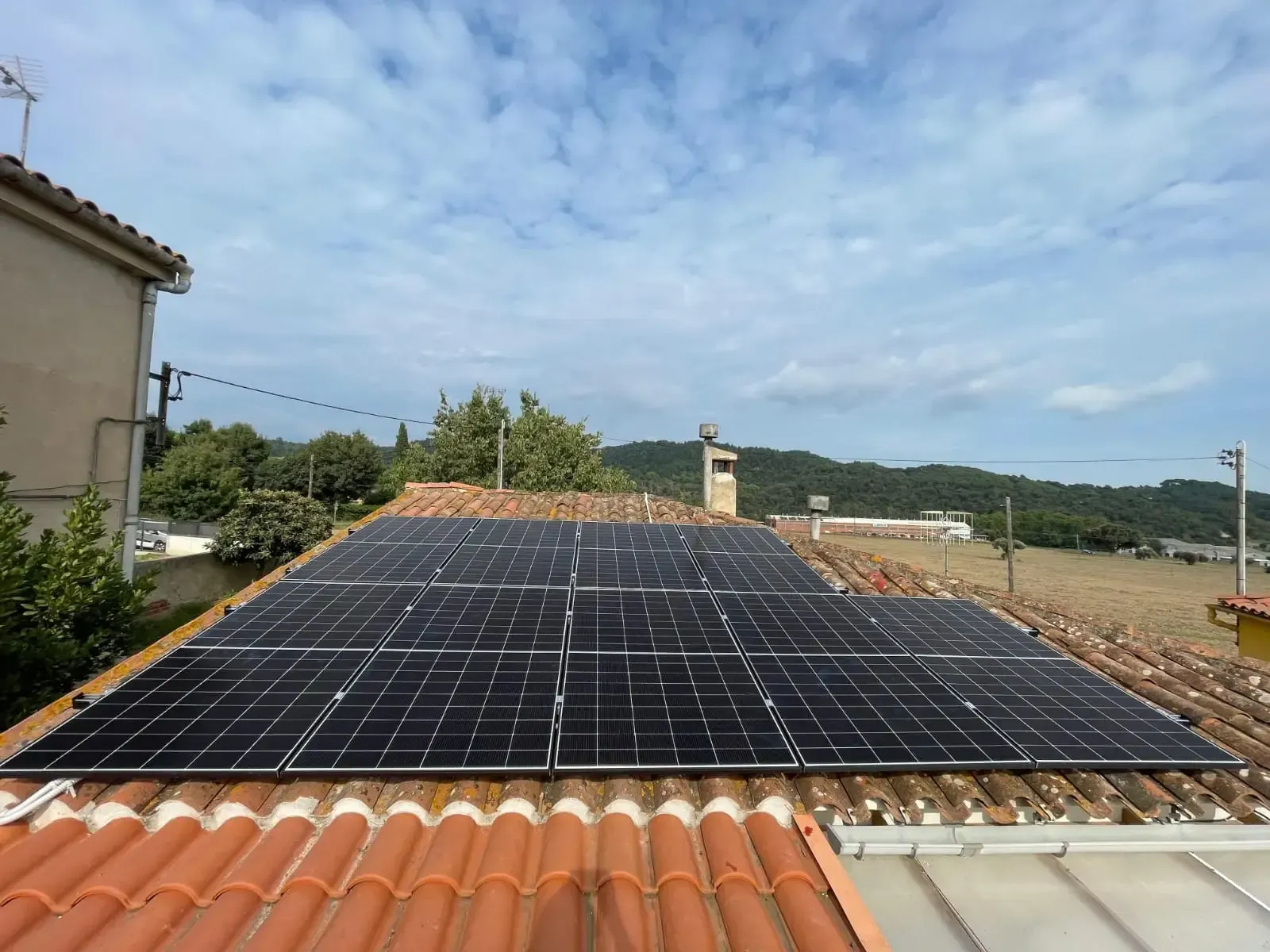 Paneles solares en la azotea bajo un cielo azul, con tejas de terracota y un paisaje rural a lo lejos.