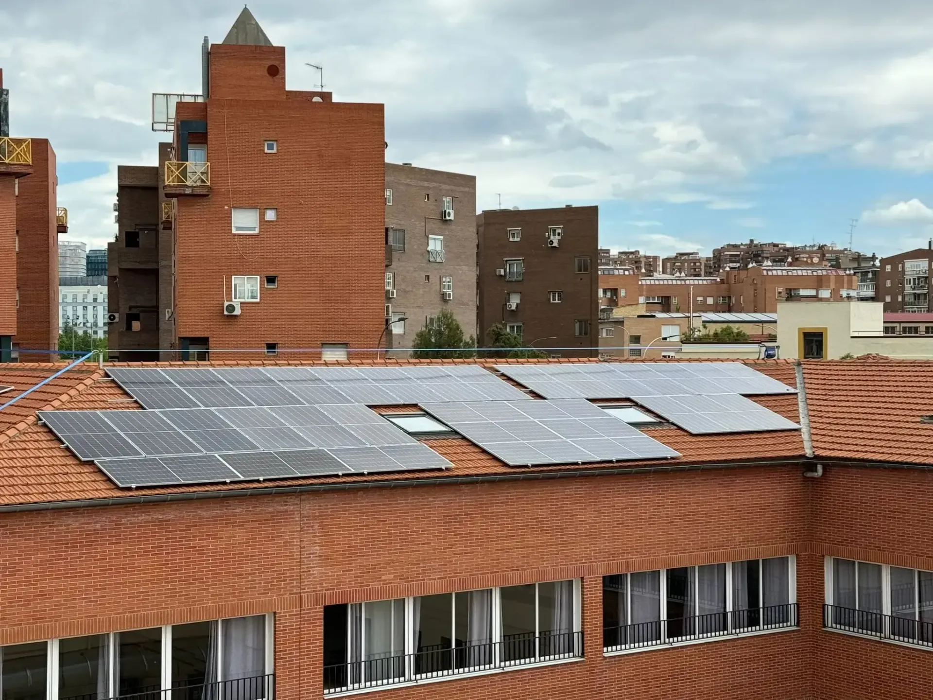 Paneles solares en el tejado de un edificio de ladrillo en un barrio urbano bajo un cielo nublado.