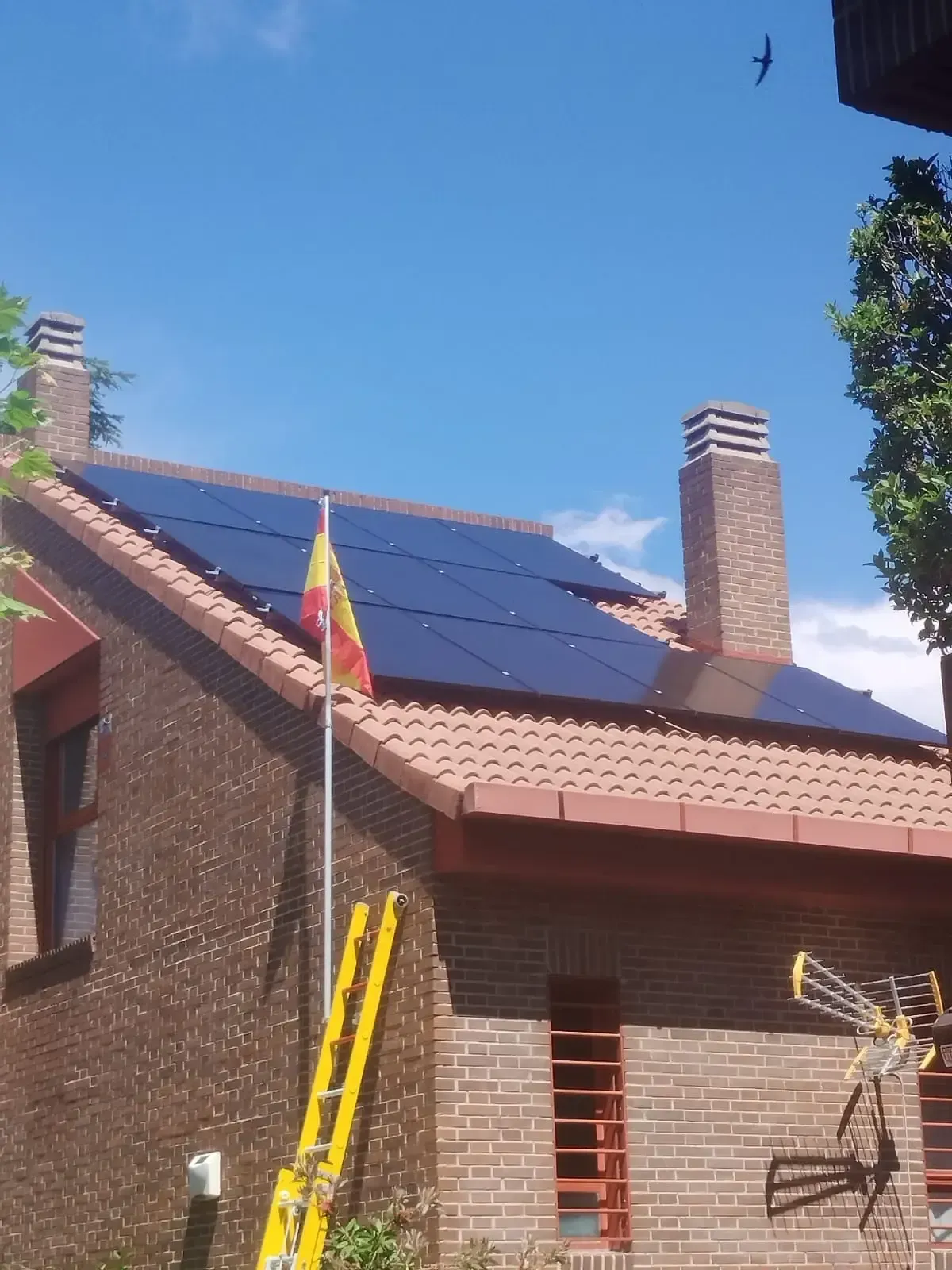 Una casa de ladrillo con paneles solares en el tejado, una escalera amarilla y un mástil bajo un cielo azul.