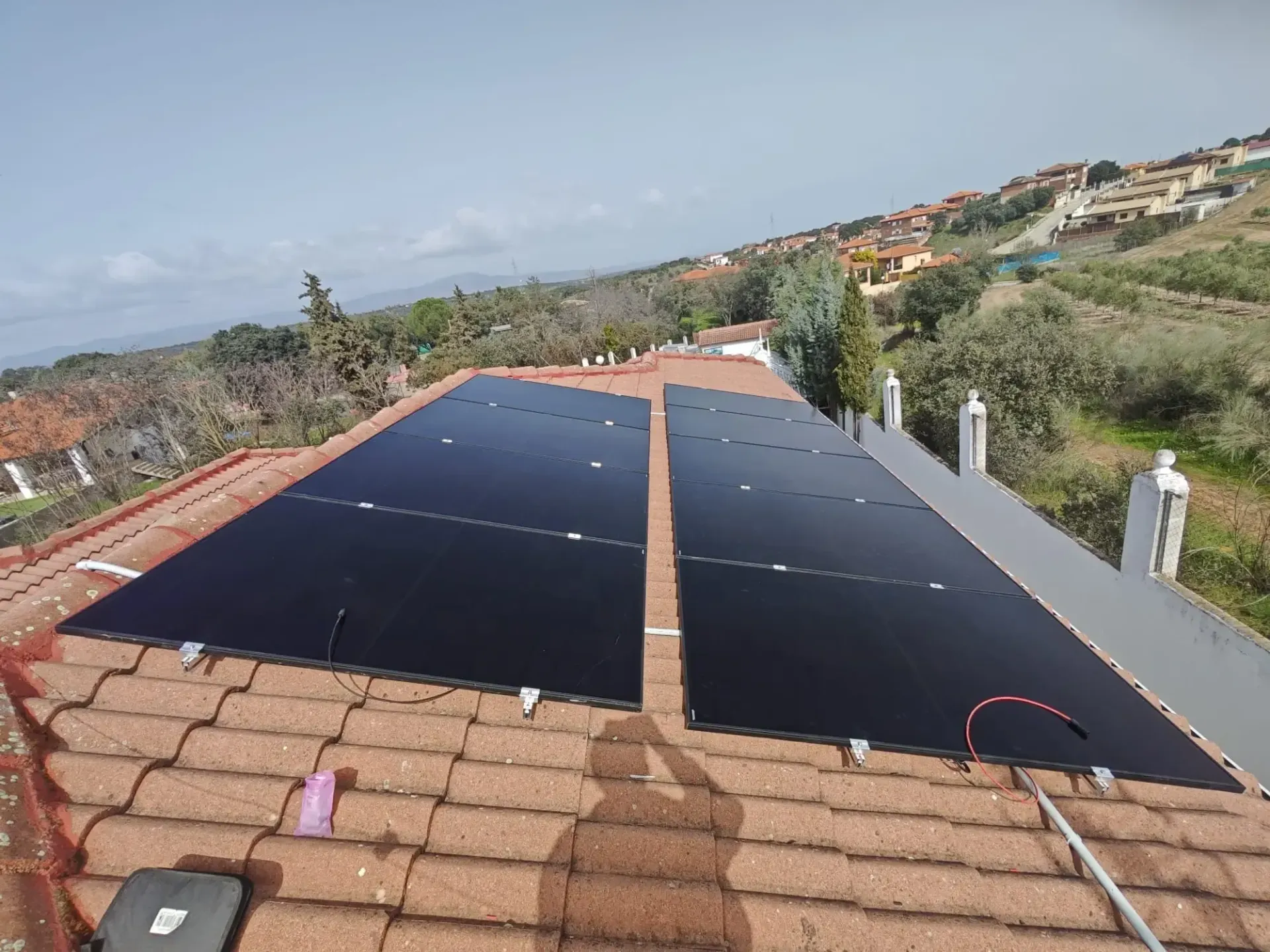 Paneles solares instalados en un tejado de tejas rojas con vistas a un barrio en la ladera de una colina bajo un cielo despejado.